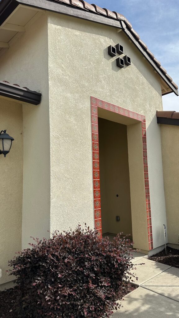 Residential exterior entryway of a house in El Dorado Hills, CA, featuring textured beige walls, decorative red trim around the doorway, modern light fixtures, and landscaped with purple foliage.